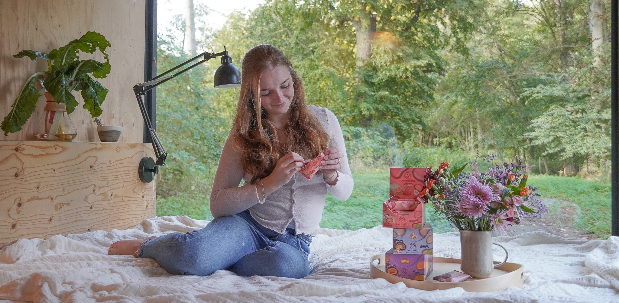 Eine Frau sitzt lächelnd auf einem Bett und öffnet eine Packung Cake Bar Strawberry Cheesecake von coconilla. Neben ihr steht ein Tablett mit einer Vase voller bunter Blumen und mehreren Schachteln Coconilla-Riegeln und Cream Bites. Im Hintergrund ist eine große Fensterfront zu sehen, durch die eine grüne Waldlandschaft sichtbar ist.